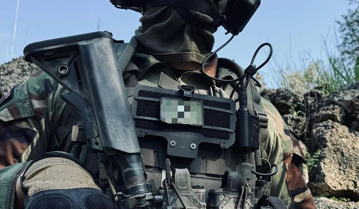 Close-up of a chest-mounted Nav-Board holder on a tactical operator's plate carrier, with a blurred tag patch.