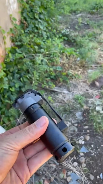 Hand holding Blank Kit training grenade with grey handle outdoors.