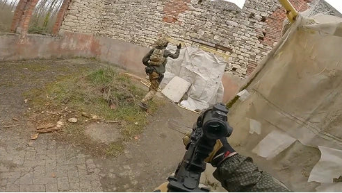 First-person perspective of an airsoft player holding a rifle, watching a teammate run past a ruined stone building during a CQB milsim game.